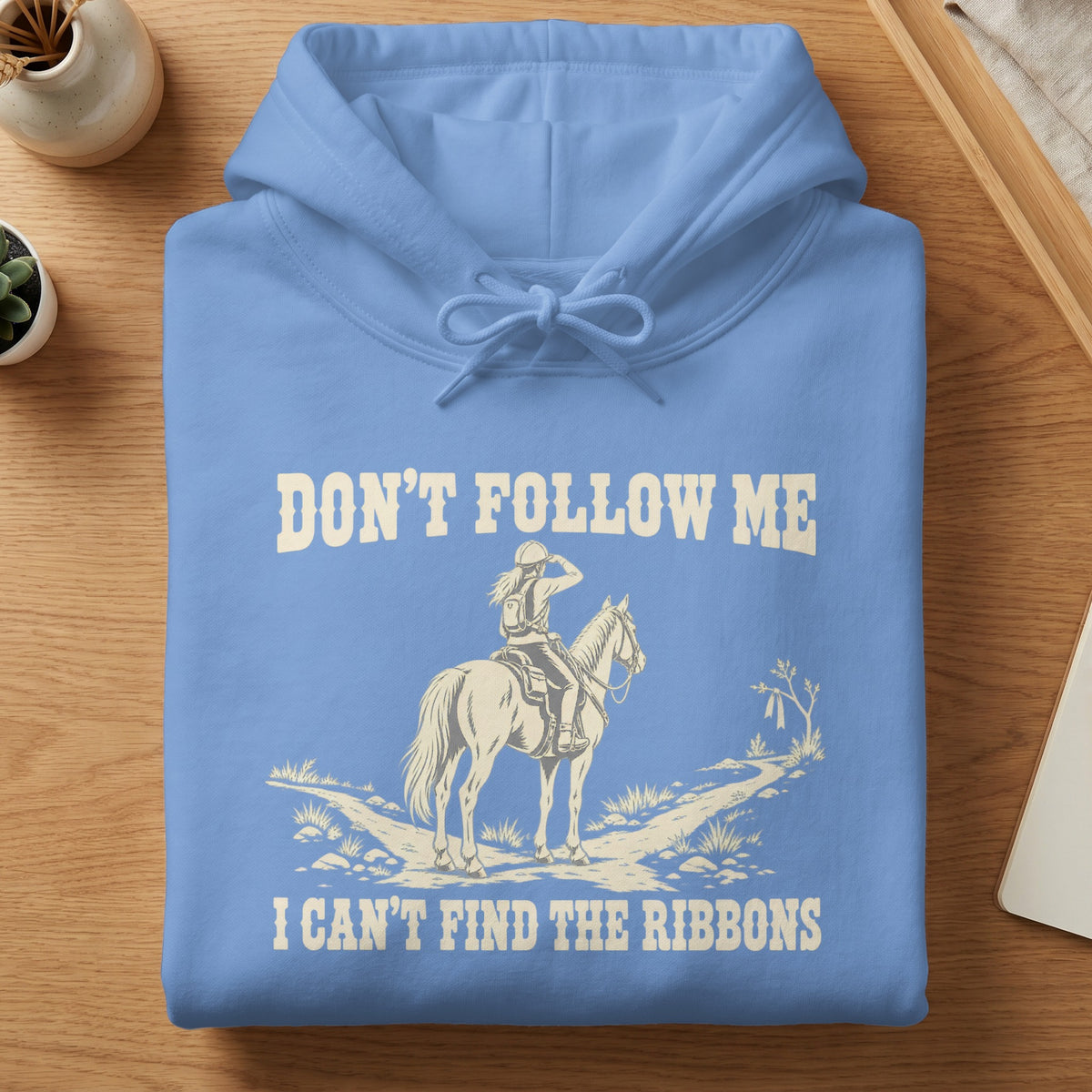 A folded blue hooded sweatshirt with white text reading "DON'T FOLLOW ME I CAN'T FIND THE RIBBONS" above and below an illustration of a horseback rider on a path.