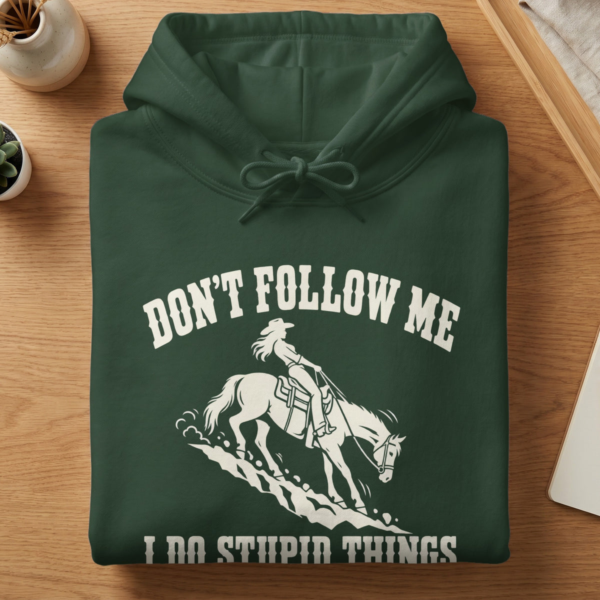 A folded dark green hoodie with white graphic text reading DON'T FOLLOW ME I DO STUPID THINGS featuring a cowgirl riding a horse downhill.