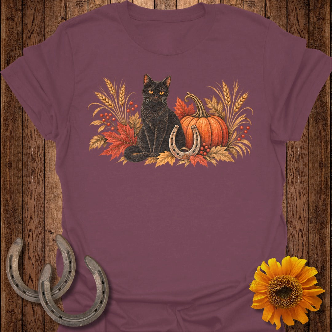 Black cat with pumpkin, horseshoe, and autumn leaves graphic on a maroon t-shirt.