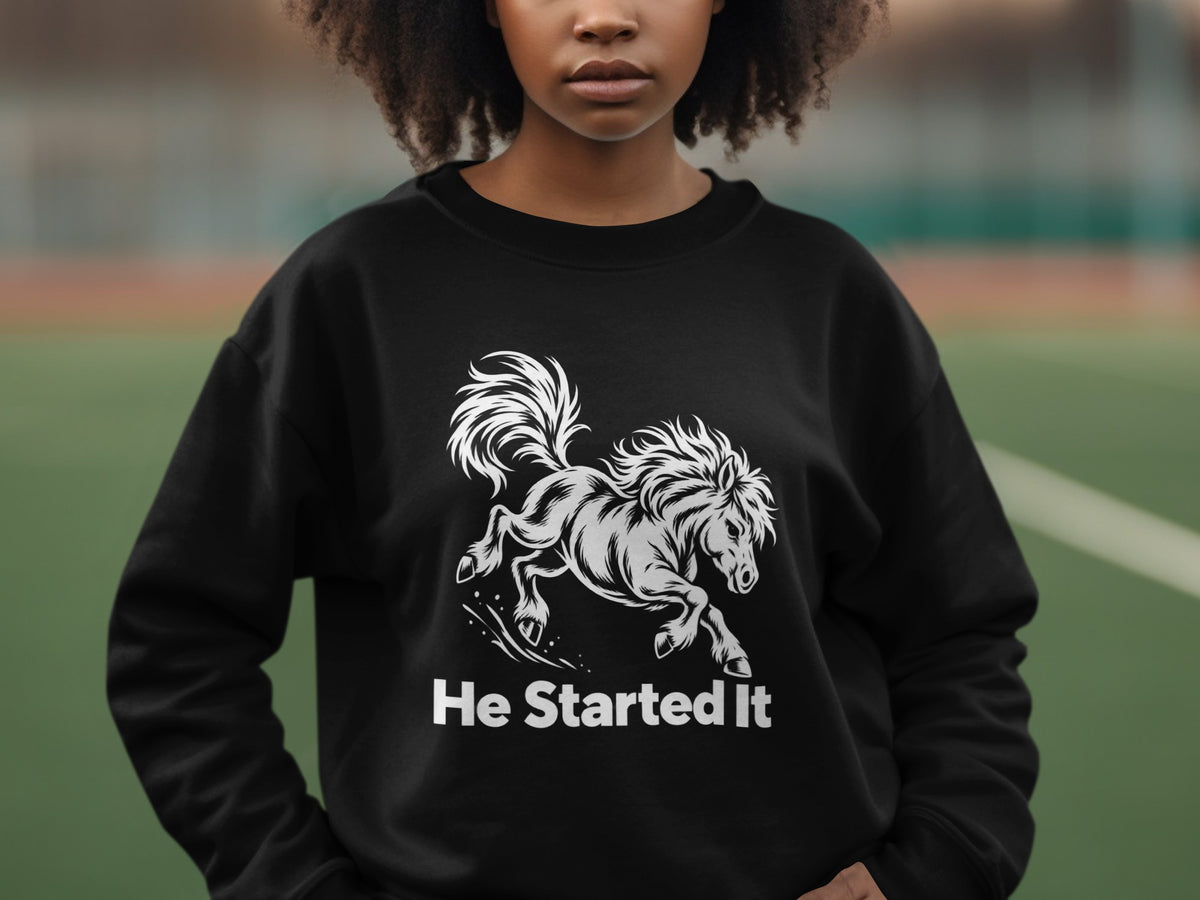 A person wearing a black crewneck sweatshirt featuring a white graphic of a running horse and the text He Started It.