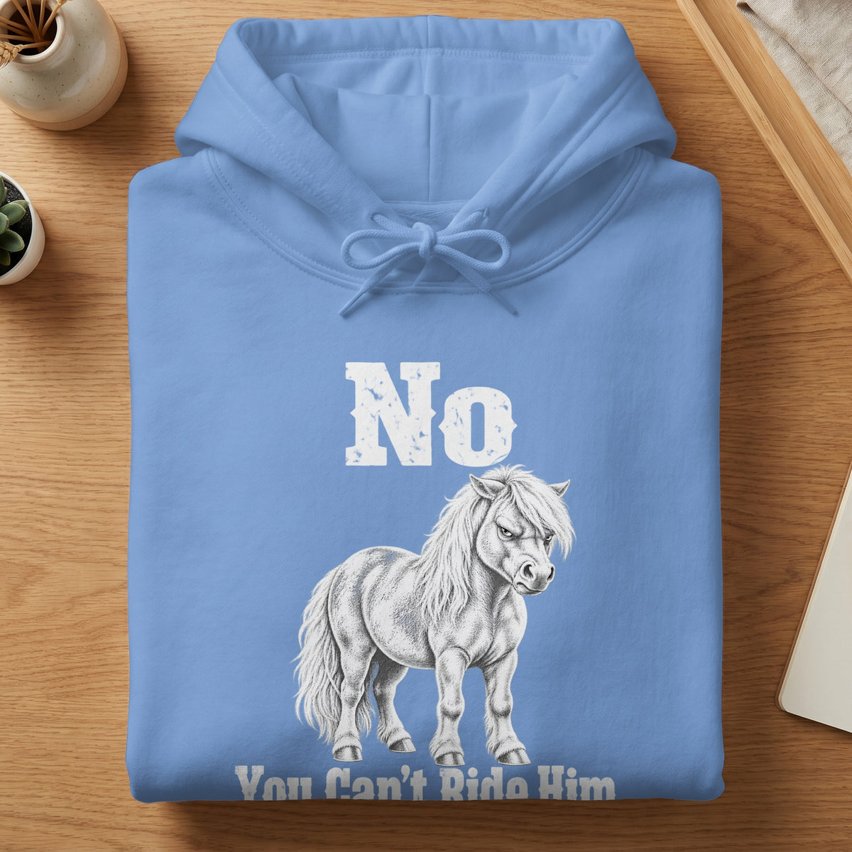 A folded blue hoodie featuring white text that reads "No" and "You Can't Ride Him" alongside a sketched, grumpy white pony.