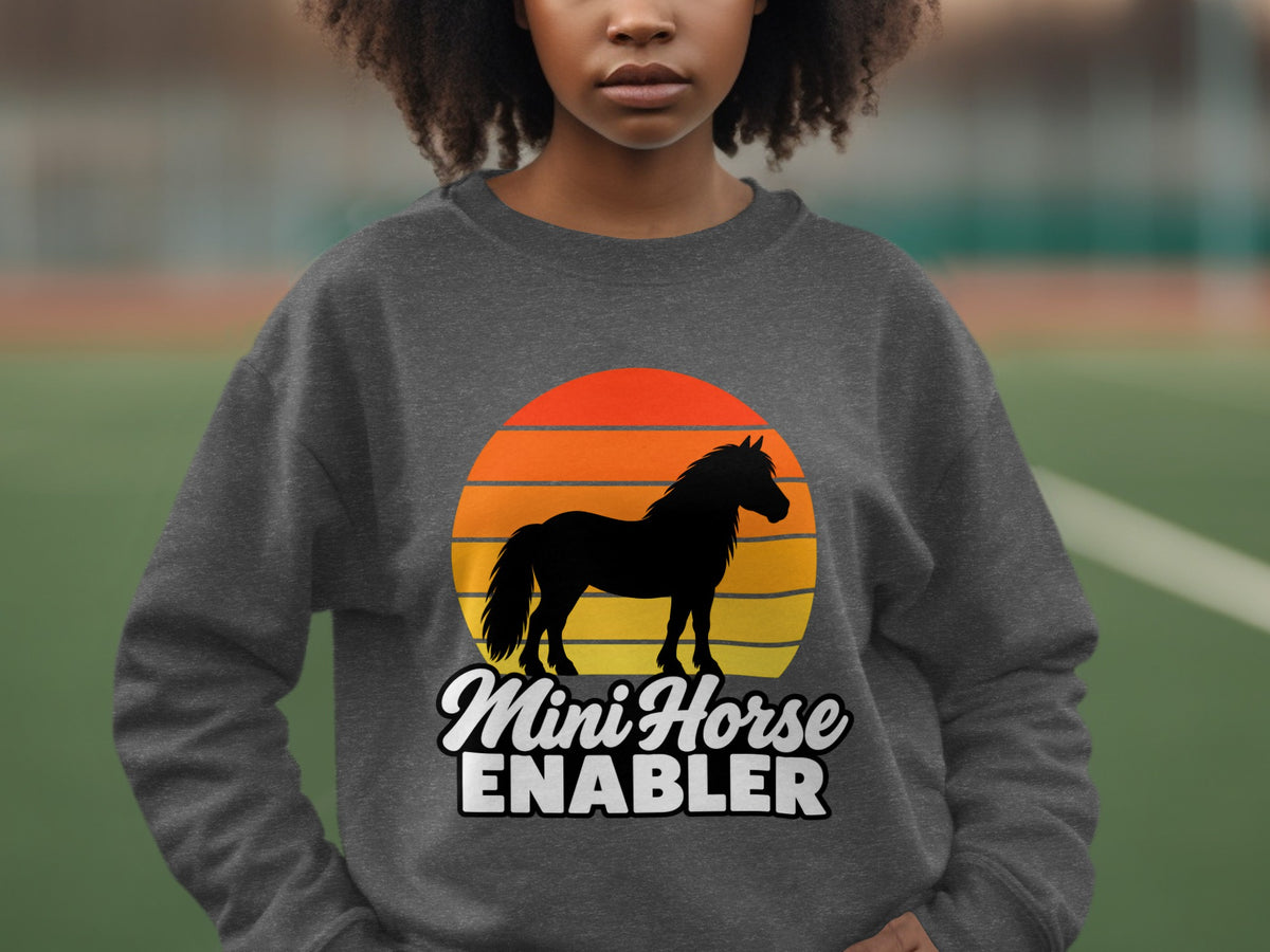 A person wearing a dark gray sweatshirt featuring a retro sunset graphic and the text MINI HORSE ENABLER.