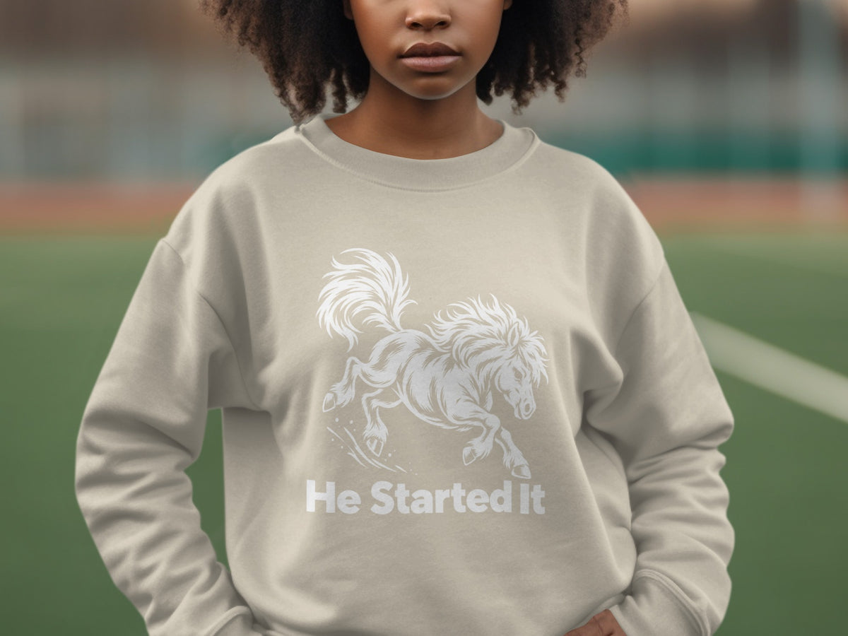 A person wearing a beige crewneck sweatshirt featuring a white running horse graphic and the text He Started It.