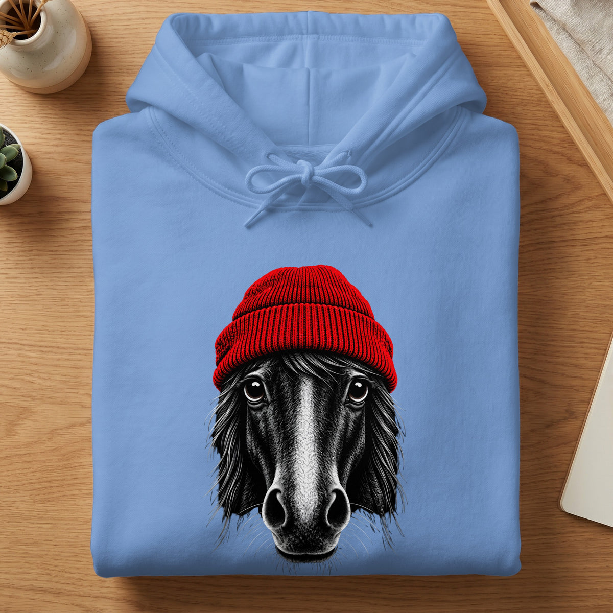 Folded light blue hooded sweatshirt featuring a graphic of a black horse wearing a red knit beanie.