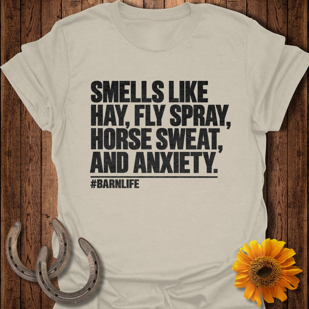 Light beige t-shirt with black text reading "SMELLS LIKE HAY, FLY SPRAY, HORSE SWEAT, AND ANXIETY. #BARNLIFE" on a wooden background with two horseshoes and a sunflower.