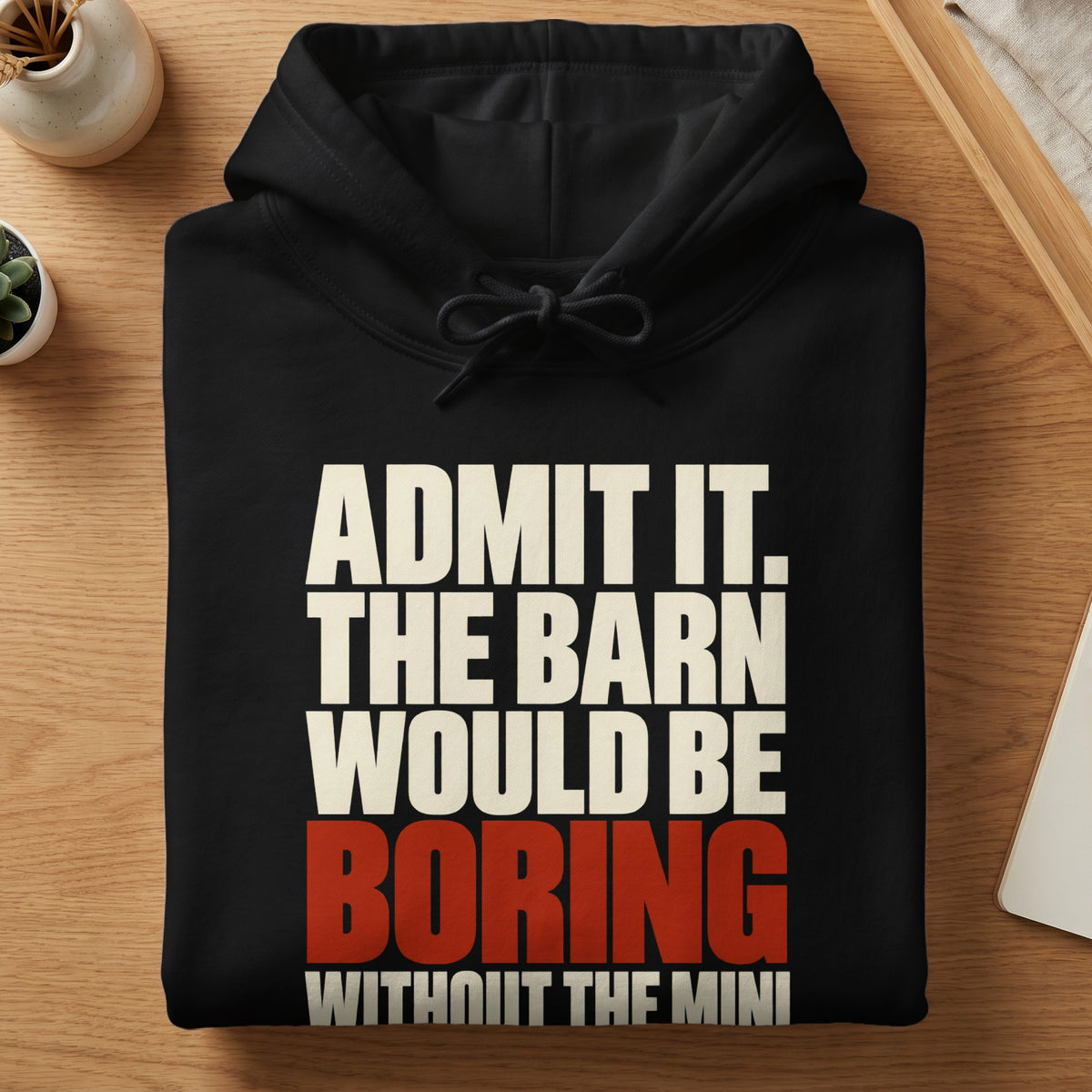 Folded black hoodie with white and red text reading ADMIT IT, THE BARN WOULD BE BORING WITHOUT THE MINI resting on a wooden surface.