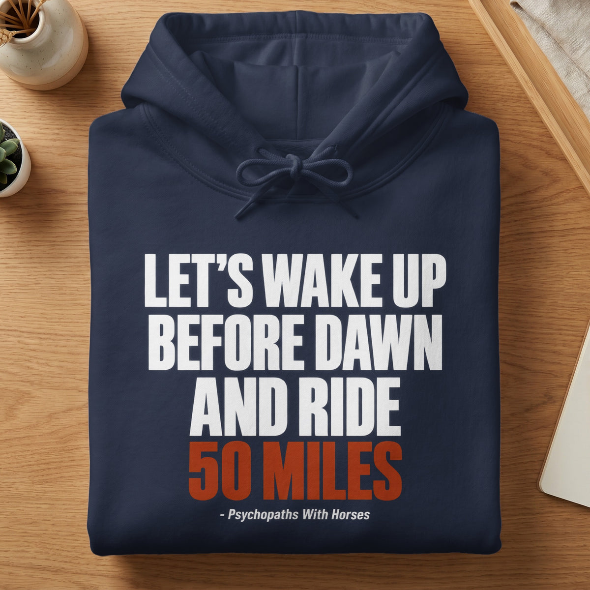 A folded navy blue hooded sweatshirt with white and orange text that reads "LET'S WAKE UP BEFORE DAWN AND RIDE 50 MILES - Psychopaths With Horses".