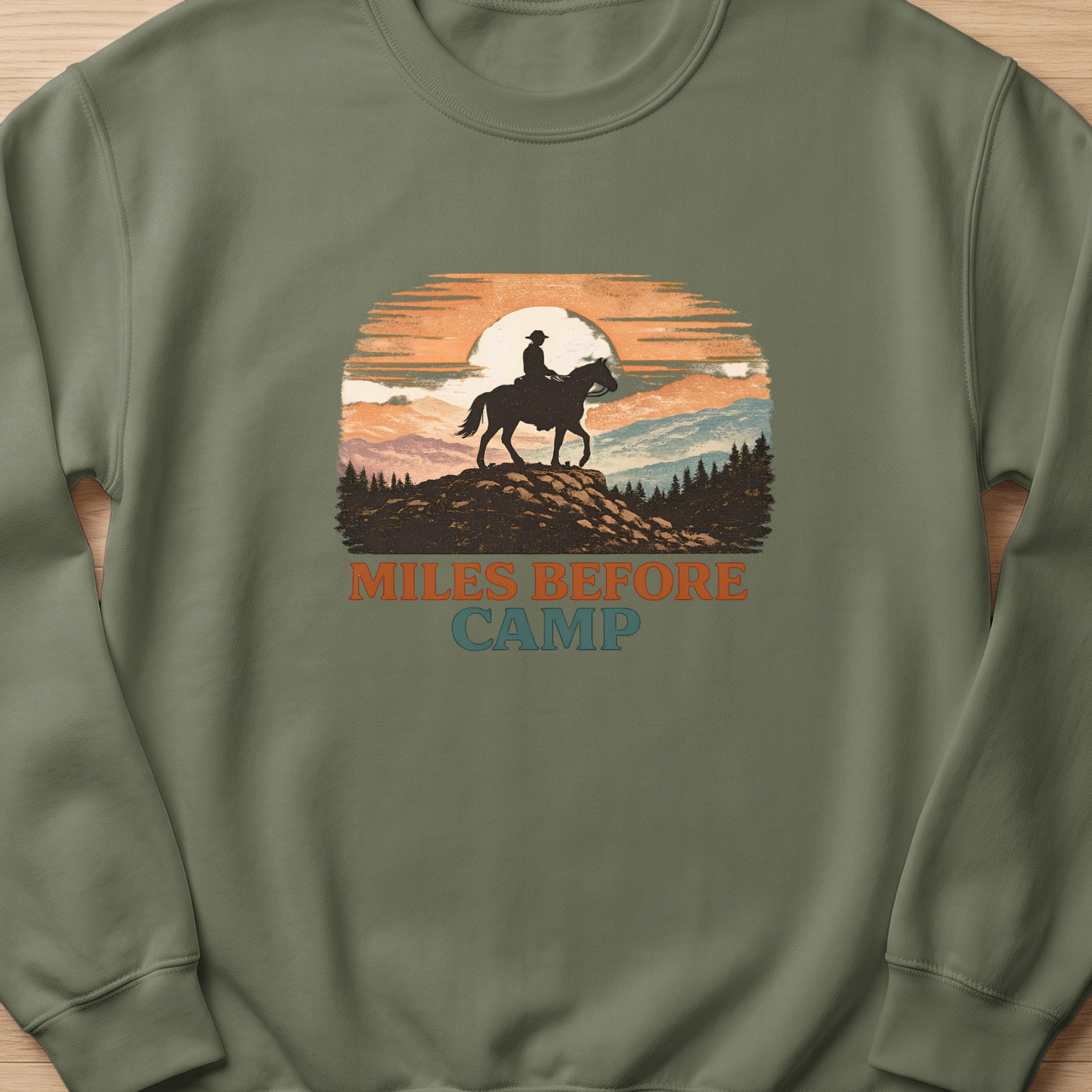 Crewneck sweatshirt with a graphic of a cowboy on horseback at sunset and text "Miles Before Camp".