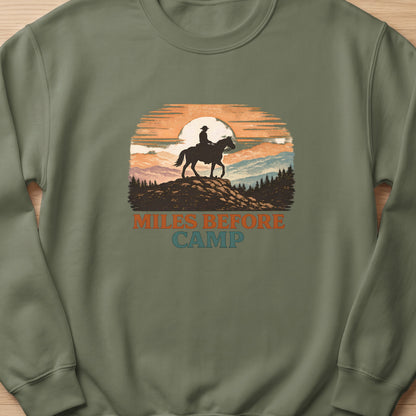 Crewneck sweatshirt with a graphic of a cowboy on horseback at sunset and text "Miles Before Camp".