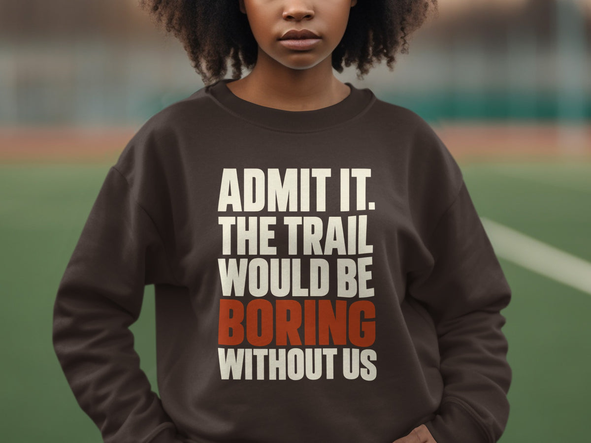 A person wears a dark brown crewneck sweatshirt featuring white and orange text reading, "ADMIT IT. THE TRAIL WOULD BE BORING WITHOUT US."