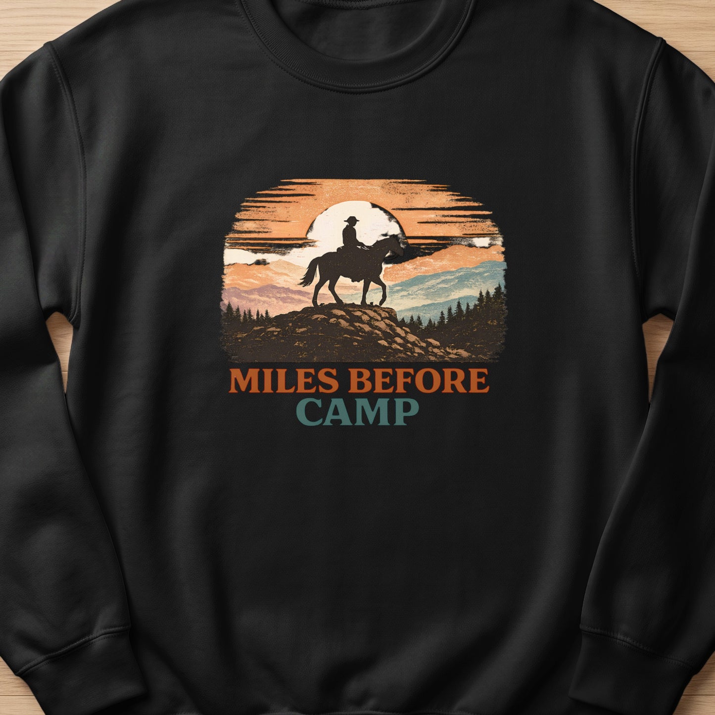 Black sweatshirt with "Miles Before Camp" text and a cowboy on horseback at sunset graphic.