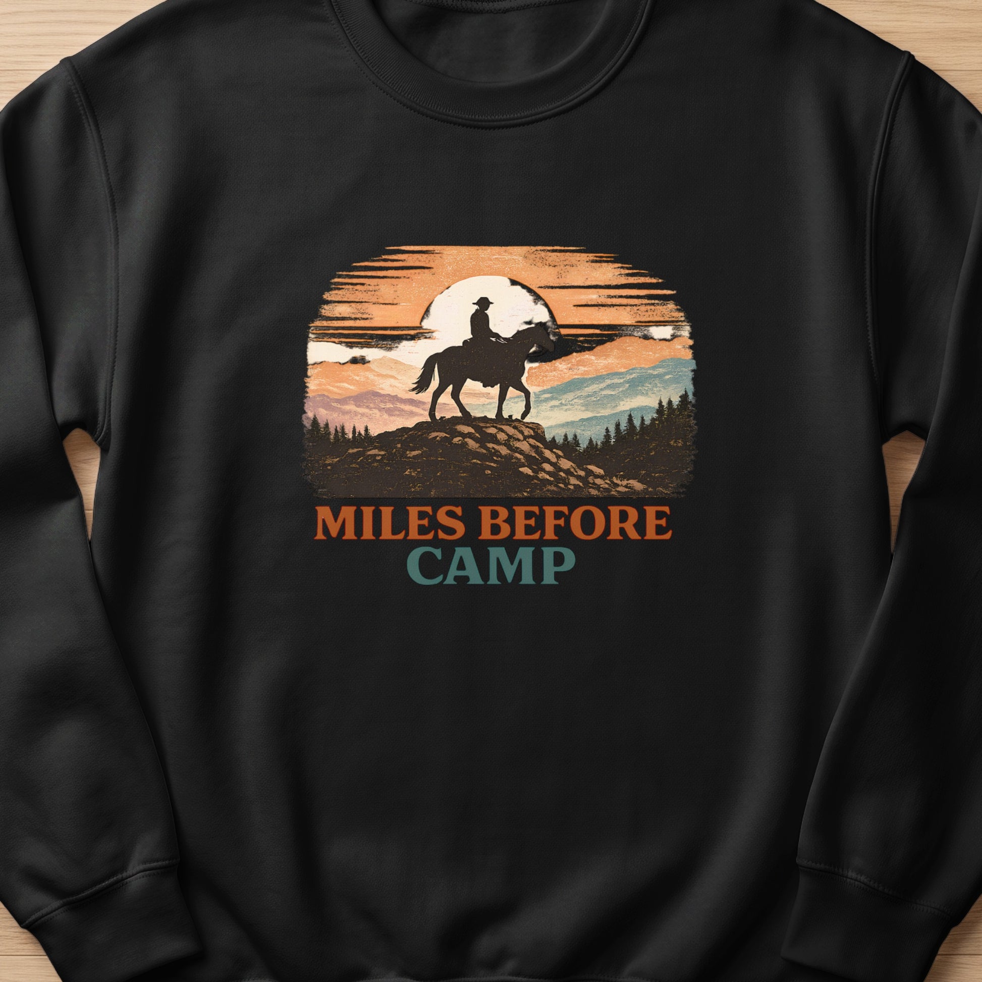 Black sweatshirt with "Miles Before Camp" text and a cowboy on horseback at sunset graphic.