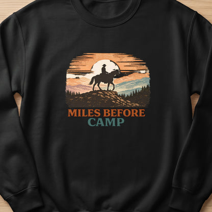 Black sweatshirt with "Miles Before Camp" text and a cowboy on horseback at sunset graphic.