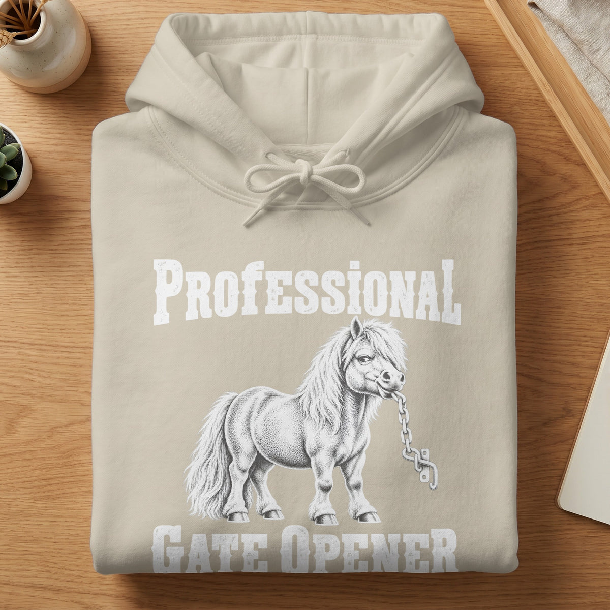 Folded beige hoodie on a wooden table, featuring white text reading Professional Gate Opener with a pony biting a chain.