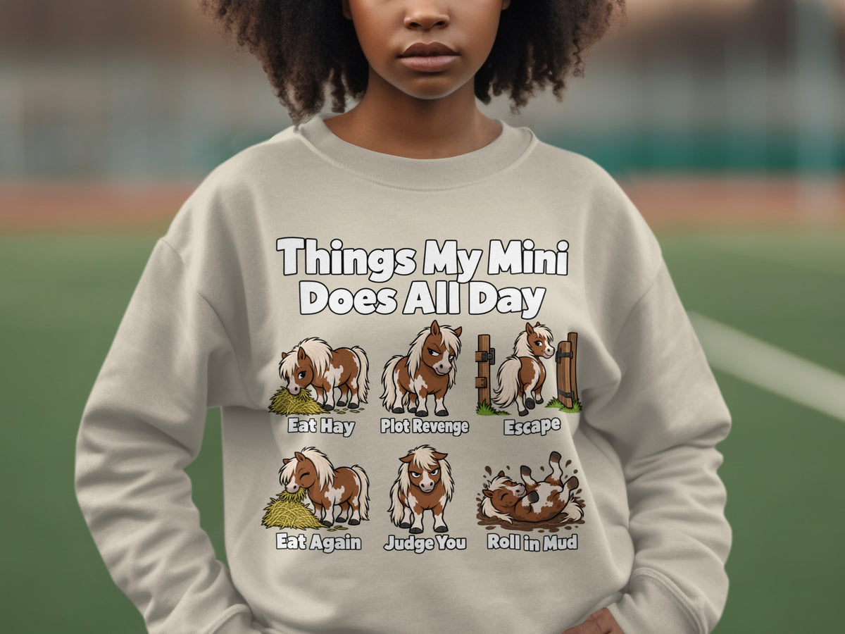 A person wearing a beige crewneck sweatshirt printed with cartoons and text about miniature horses.