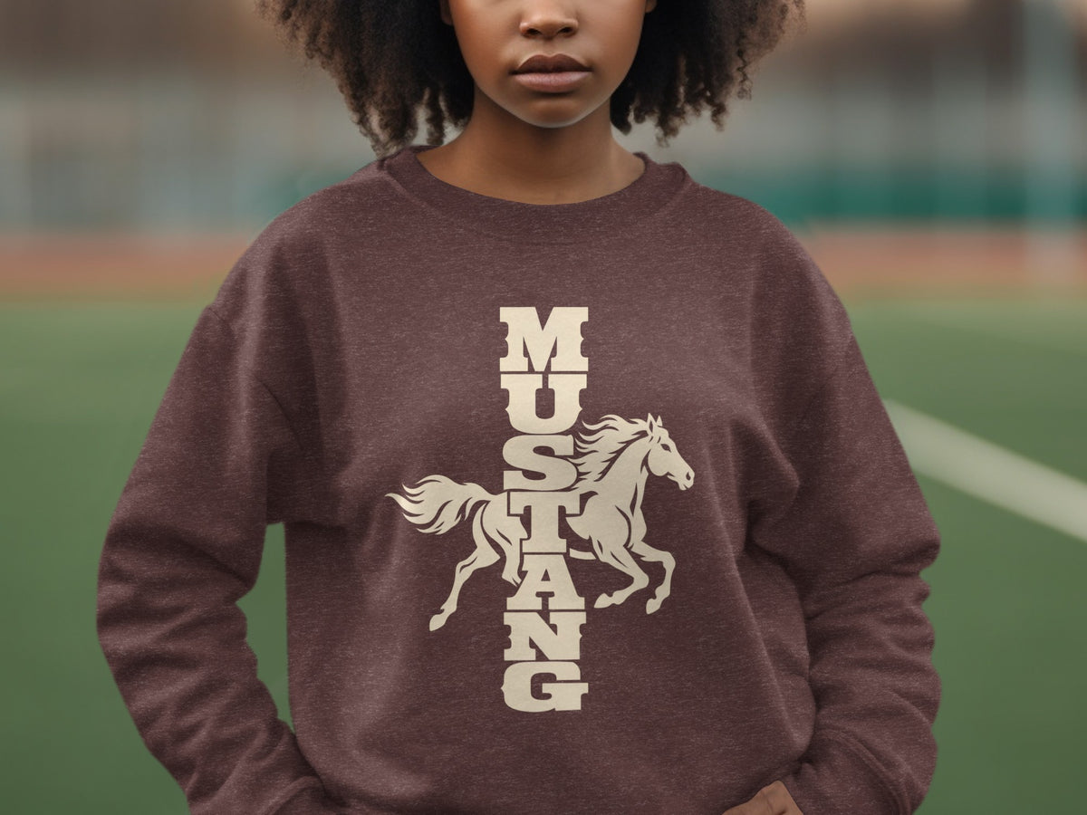 A person wearing a brown crewneck sweatshirt featuring a cream graphic of a running horse and vertical Mustang text.