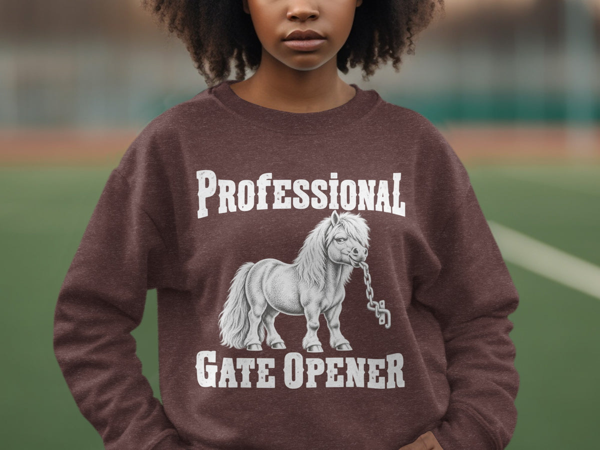 A person wearing a brown crewneck sweatshirt featuring a graphic of a pony holding a chain in its mouth, with text reading PROFESSIONAL GATE OPENER.