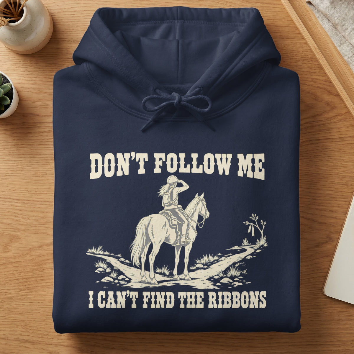 A folded dark blue hoodie featuring a white graphic of a person on a horse, with the text "DON'T FOLLOW ME I CAN'T FIND THE RIBBONS".