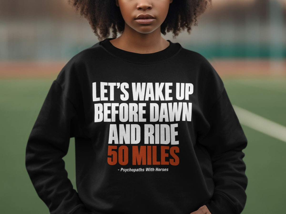 A person wearing a black crewneck sweatshirt with the text "LET'S WAKE UP BEFORE DAWN AND RIDE 50 MILES" printed in white and orange.