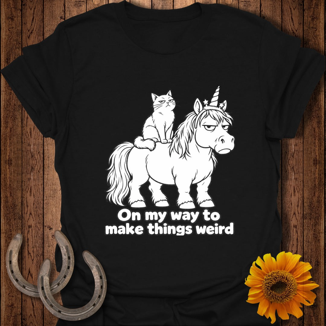A black t-shirt features a white graphic of an annoyed cat riding a grumpy unicorn, with text reading "On my way to make things weird".