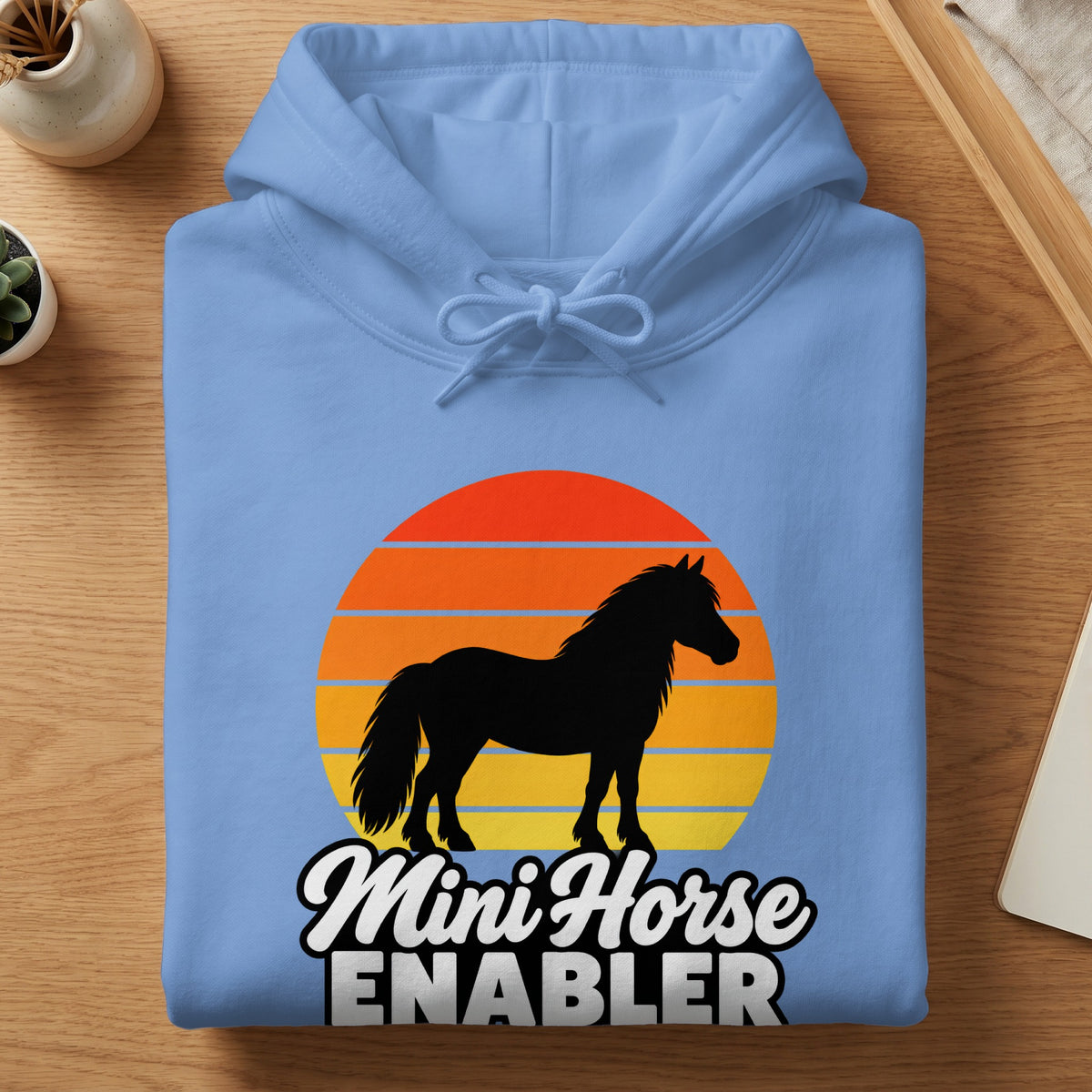 Light blue hoodie folded on a wooden surface, featuring a black silhouette of a horse against a sunset graphic with the text Mini Horse Enabler.