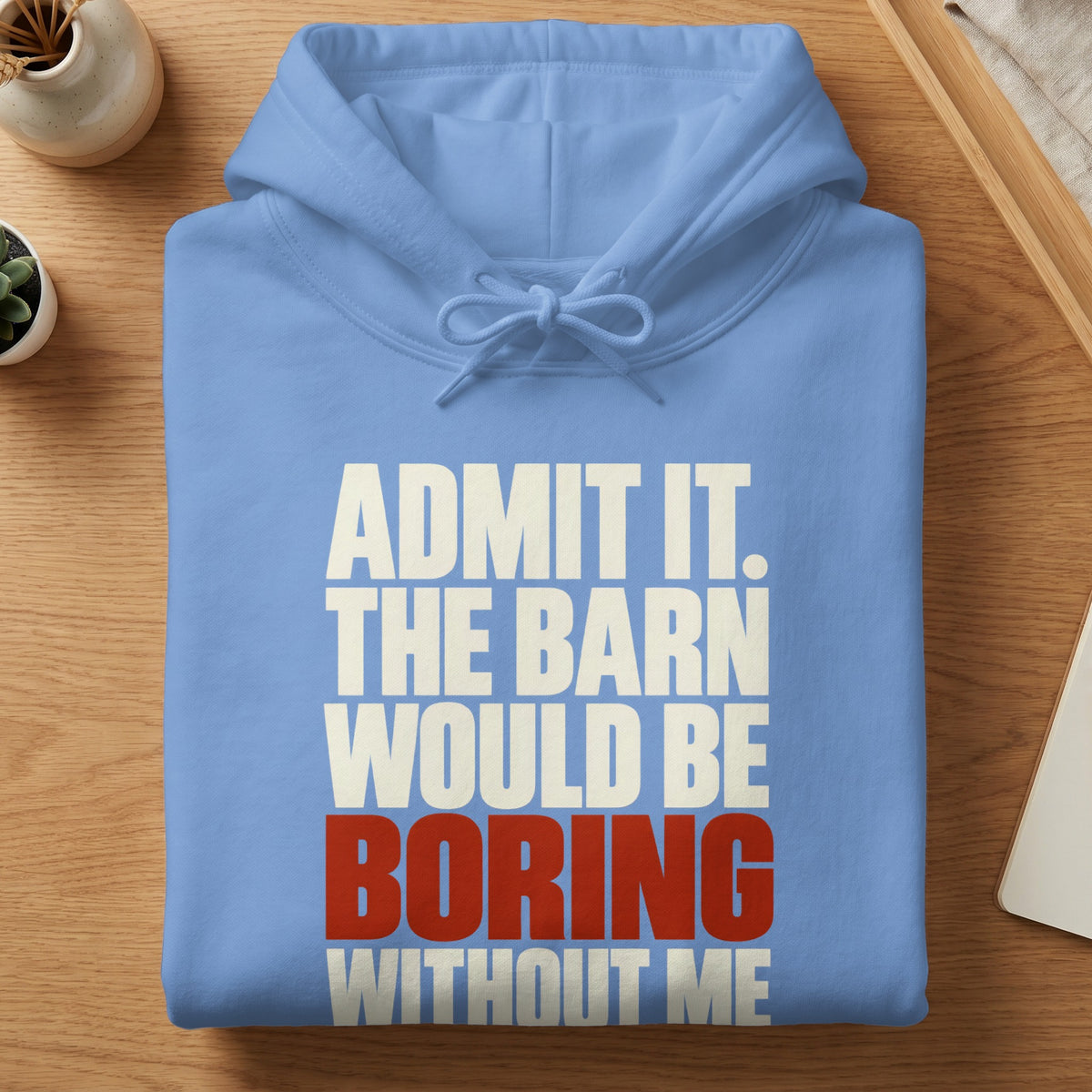 A light blue hoodie with white and red text reading ADMIT IT. THE BARN WOULD BE BORING WITHOUT ME, folded flat on a wooden surface.