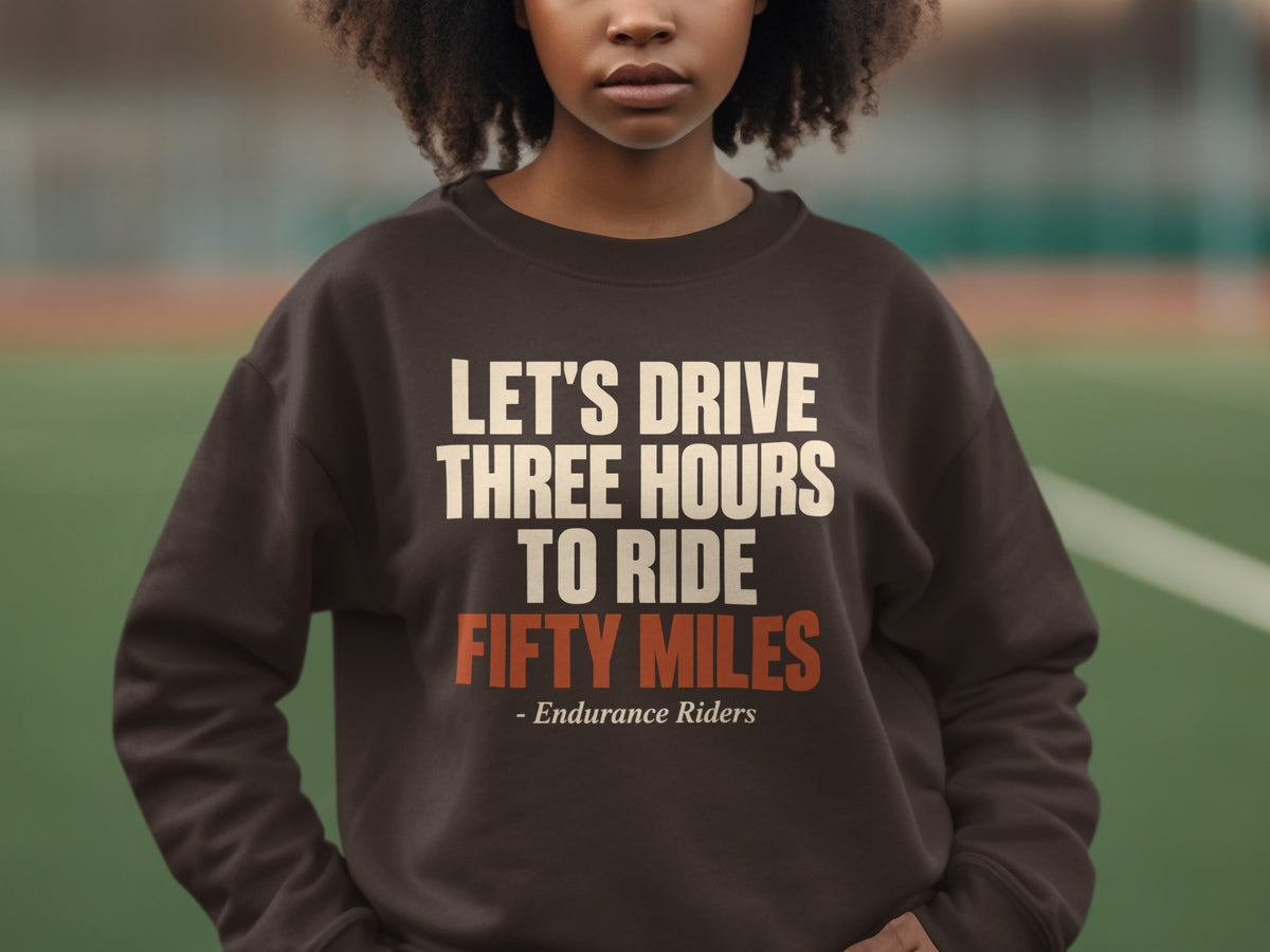 A person wearing a brown crewneck sweatshirt featuring the text Let's Drive Three Hours To Ride Fifty Miles - Endurance Riders.