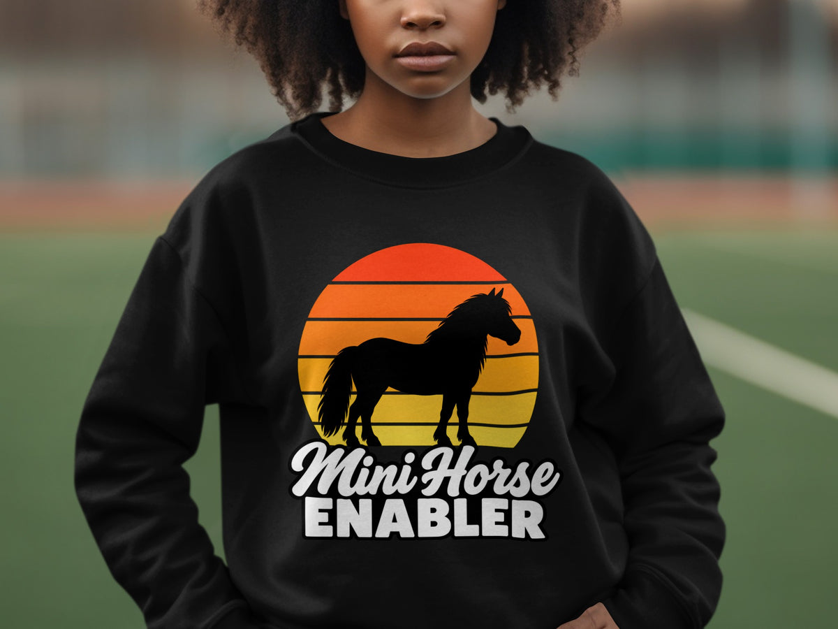 A person wearing a black crewneck sweatshirt printed with a horse silhouette, sunset graphic, and text reading Mini Horse ENABLER.