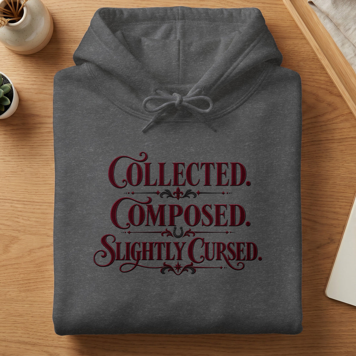 Dark grey hoodie with "Collected. Composed. Slightly Cursed." text.