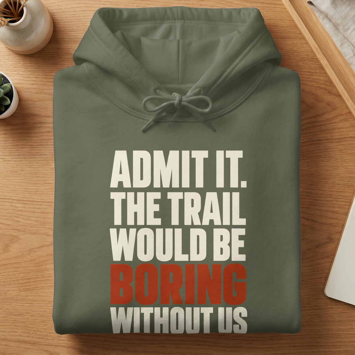 An olive green hooded sweatshirt folded on a wooden table, featuring white and red text that reads "ADMIT IT. THE TRAIL WOULD BE BORING WITHOUT US."