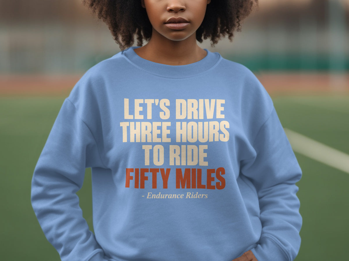 A person wears a light blue crewneck sweatshirt with the text "LET'S DRIVE THREE HOURS TO RIDE FIFTY MILES - Endurance Riders" printed on the chest.