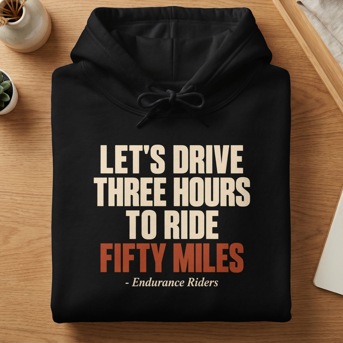 A folded black hooded sweatshirt on a wooden table features text reading, Let's Drive Three Hours to Ride Fifty Miles - Endurance Riders.