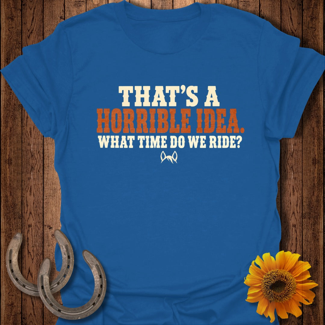 Blue crew-neck t-shirt with text saying THAT'S A HORRIBLE IDEA. WHAT TIME DO WE RIDE? on a wooden background with horseshoes and a sunflower.