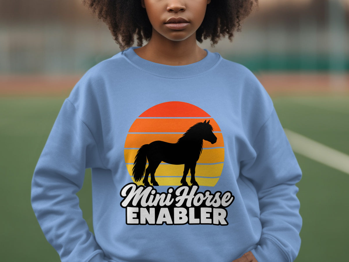 A person wears a light blue crewneck sweatshirt featuring a black horse silhouette over a sunset graphic with text reading Mini Horse Enabler.