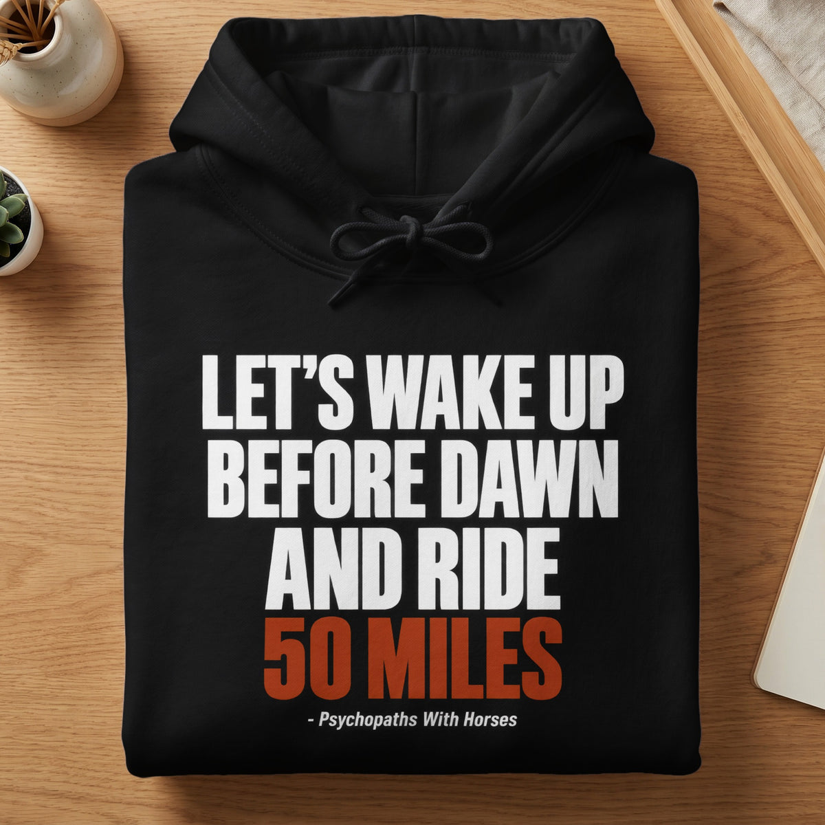 A folded black hoodie featuring white and orange text that reads Let us wake up before dawn and ride 50 miles.
