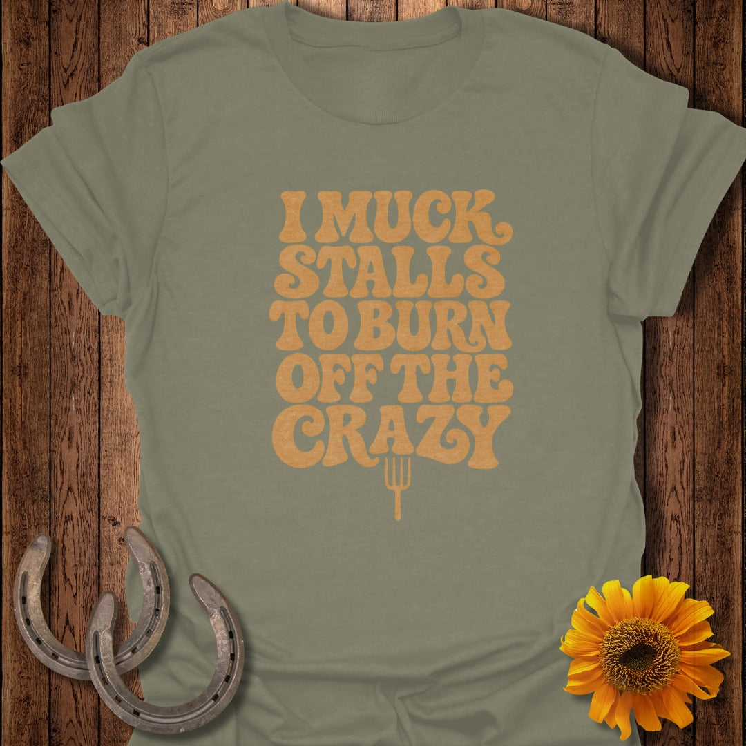 An olive green t-shirt with orange text reading I MUCK STALLS TO BURN OFF THE CRAZY and a pitchfork icon, on a wooden background with horseshoes and a sunflower.