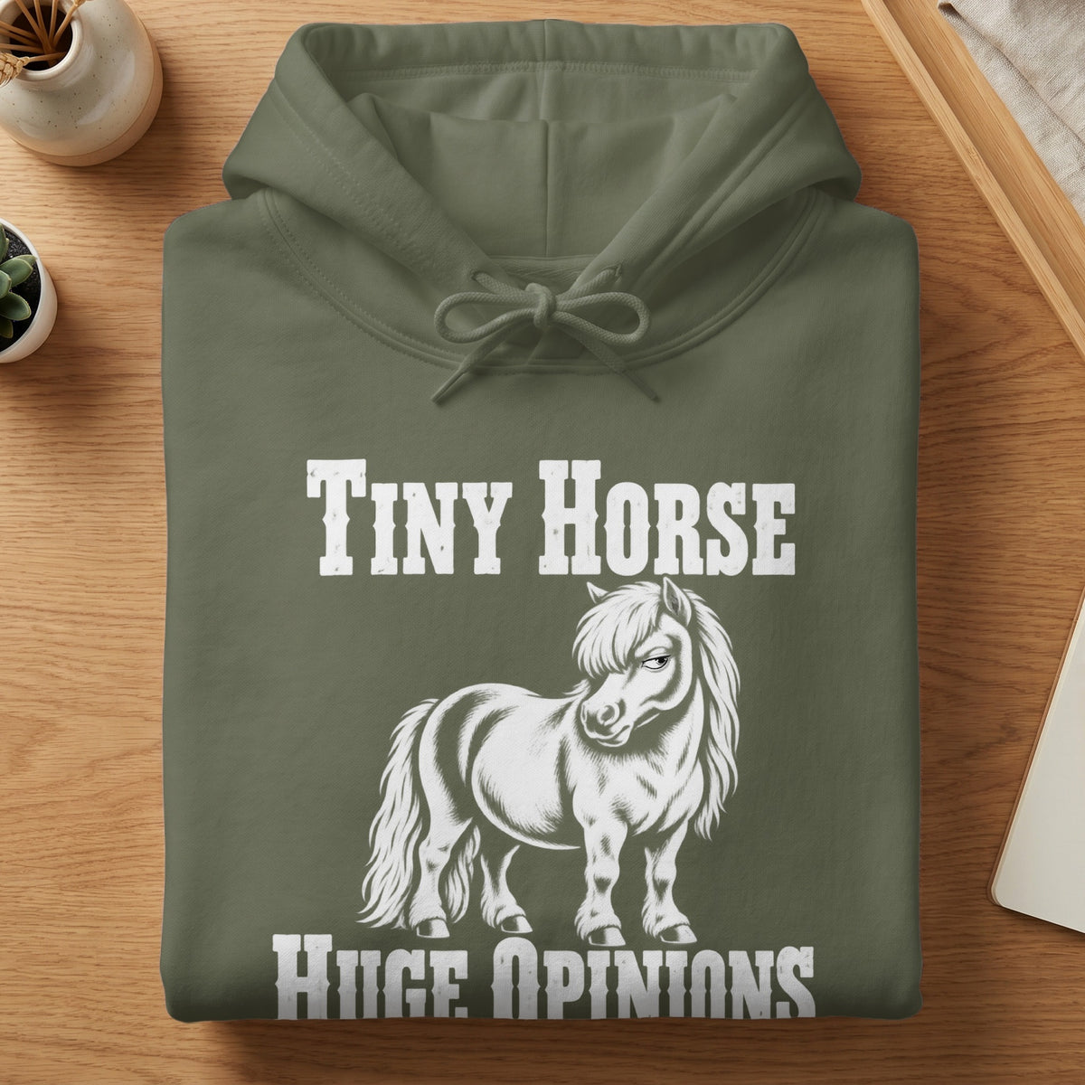 An olive green hoodie folded on a wooden table, printed with a white graphic of a grumpy pony and text reading Tiny Horse Huge Opinions.