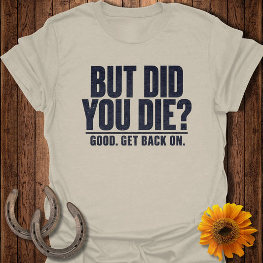 A light gray short-sleeved t-shirt features the distressed dark blue text "BUT DID YOU DIE? GOOD. GET BACK ON." against a wooden backdrop decorated with horseshoes and a sunflower.