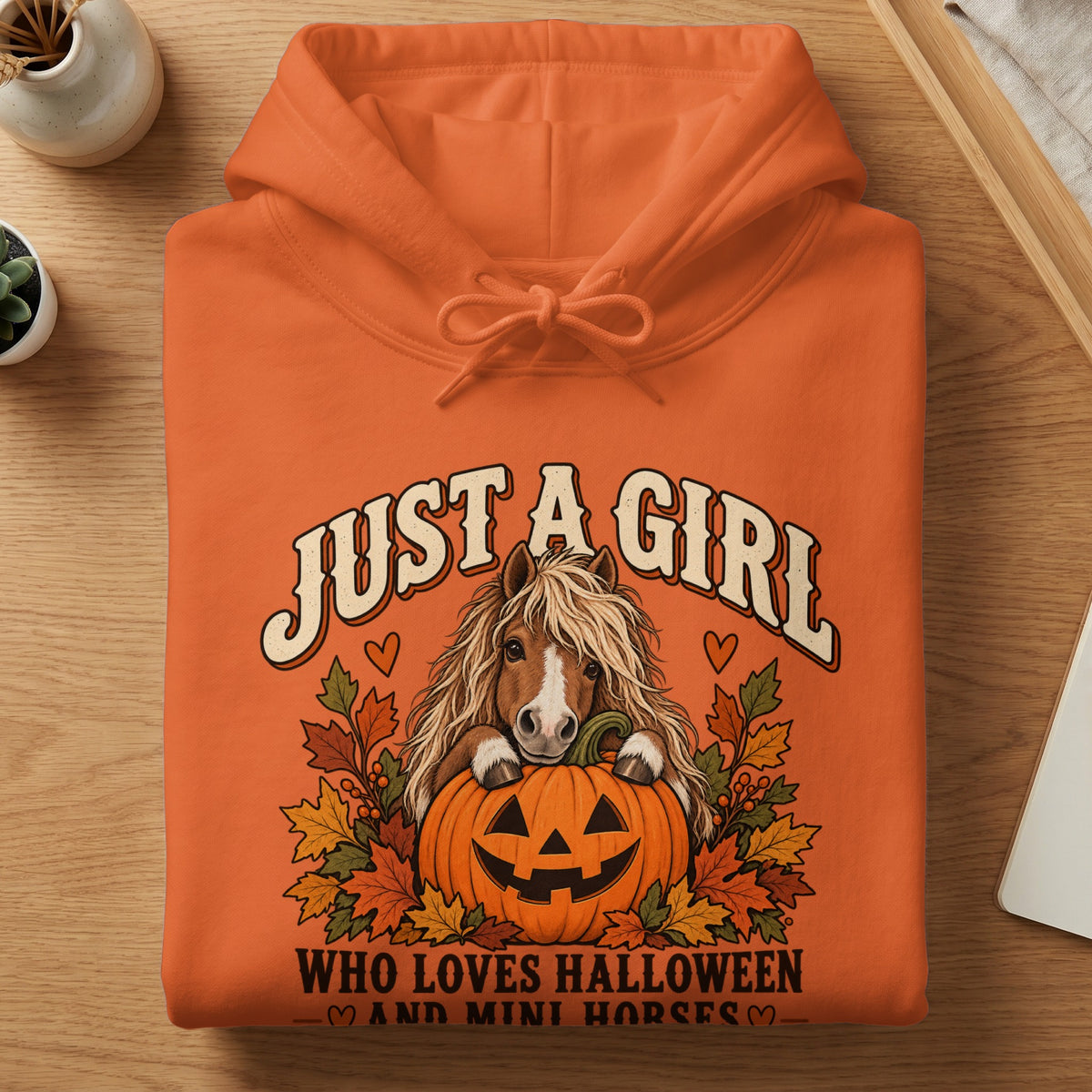 An orange hooded sweatshirt featuring a graphic of a pony behind a jack-o-lantern with autumn leaves.