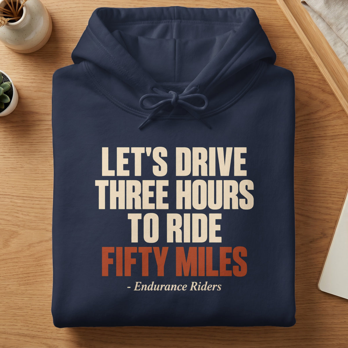Folded dark blue hoodie resting on a wooden surface, featuring the text Let's Drive Three Hours to Ride Fifty Miles Endurance Riders in white and orange.