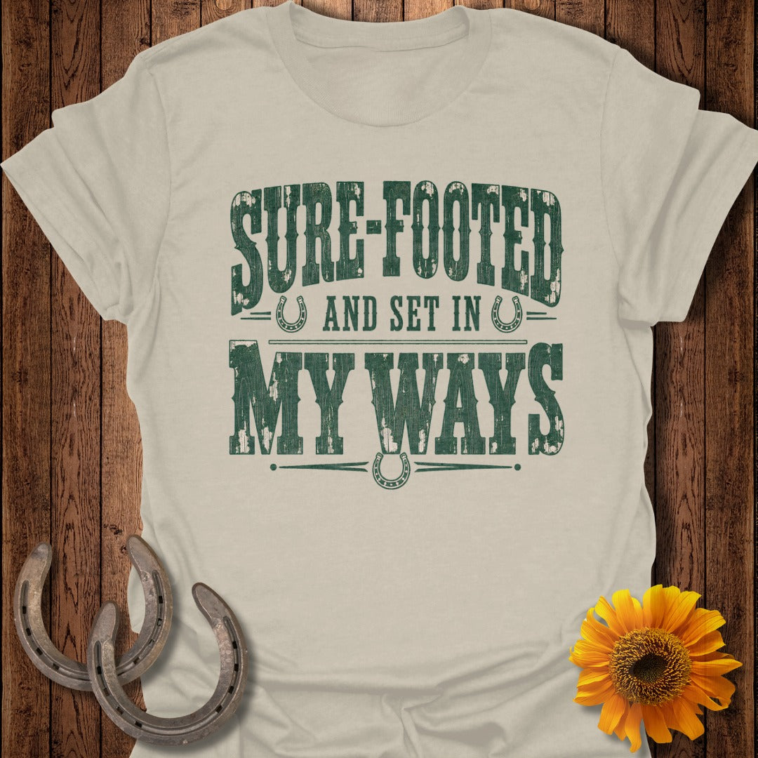 Light tan t-shirt with distressed green text reading "SURE-FOOTED AND SET IN MY WAYS".
