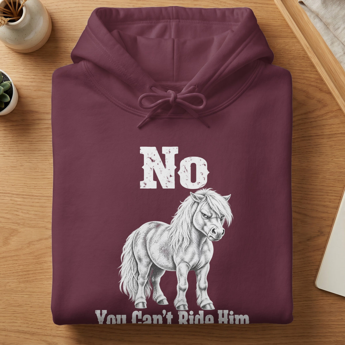 A folded maroon hooded sweatshirt featuring white text that reads "No You Can't Ride Him" alongside a sketch of a pony.