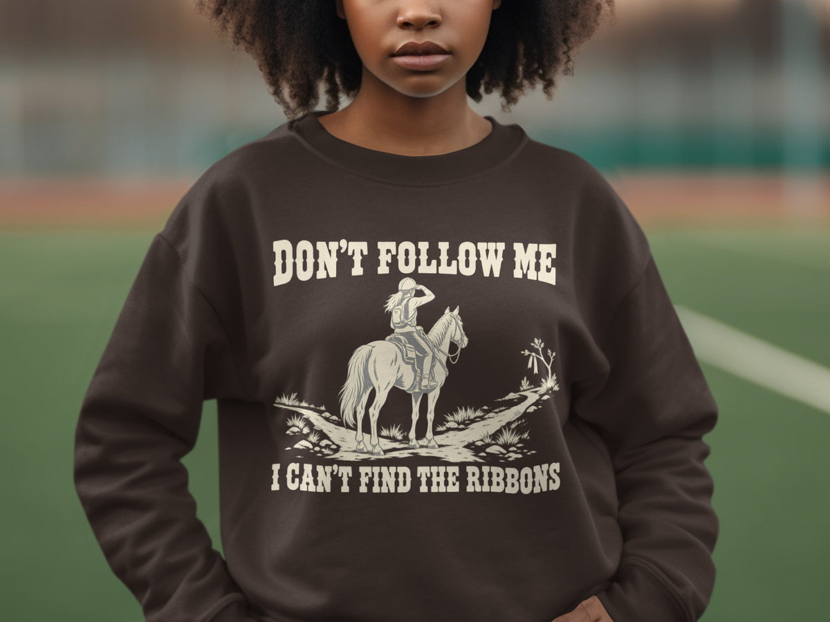 A person wearing a dark brown crewneck sweatshirt featuring an illustration of a horseback rider and the text, "DON'T FOLLOW ME I CAN'T FIND THE RIBBONS."