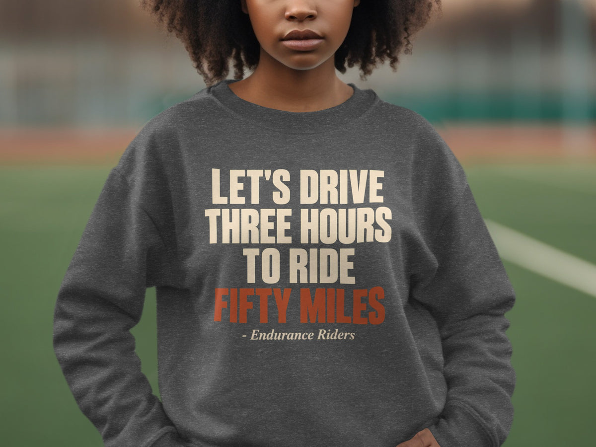 A person wearing a dark gray crewneck sweatshirt with the text "Let's Drive Three Hours To Ride Fifty Miles - Endurance Riders" printed on the chest.
