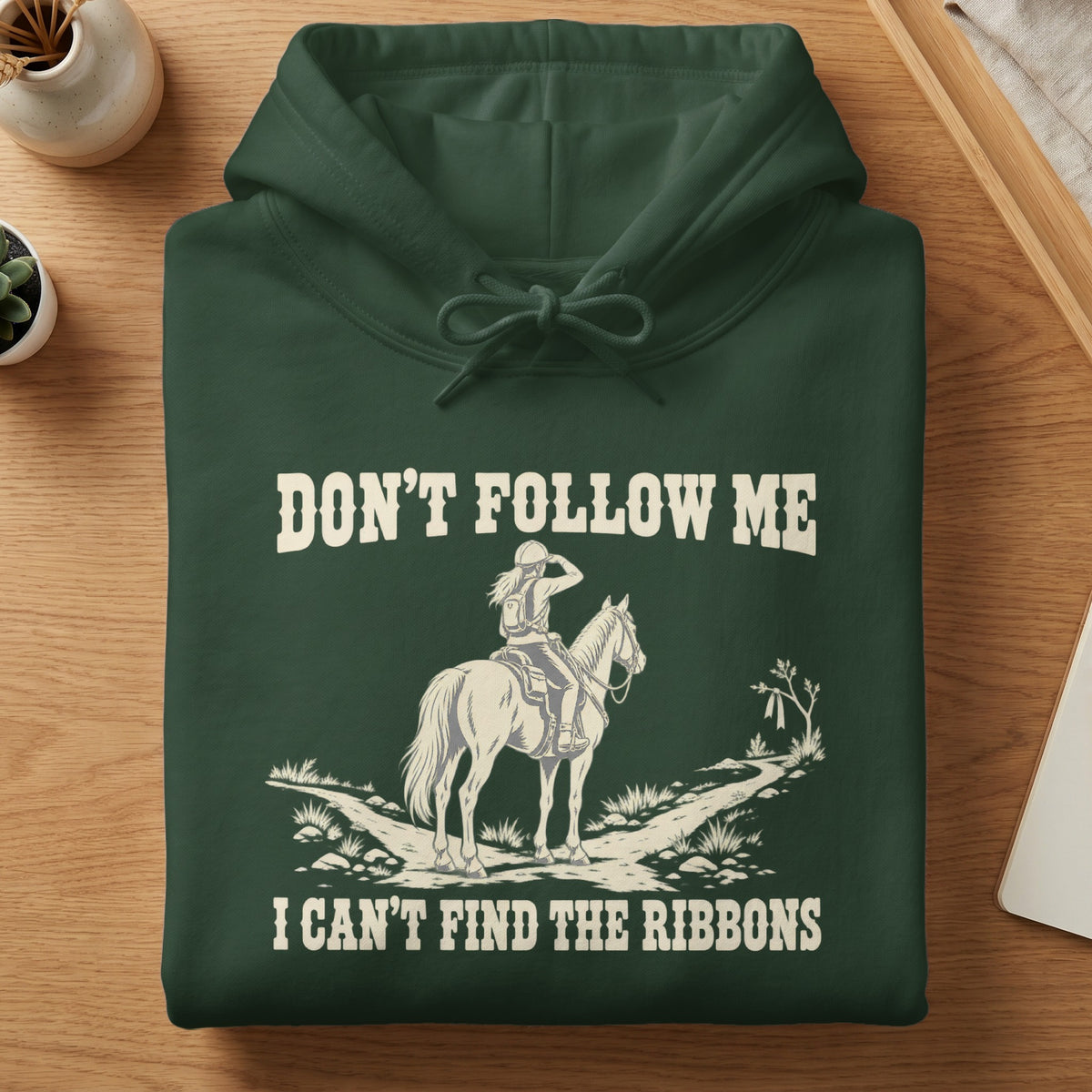 A folded dark green hoodie features a graphic of a horse and rider with the text DON'T FOLLOW ME I CAN'T FIND THE RIBBONS.