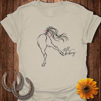 Light tan t-shirt with black line art of a horse kicking, a pink bow, and "still kicking" text.