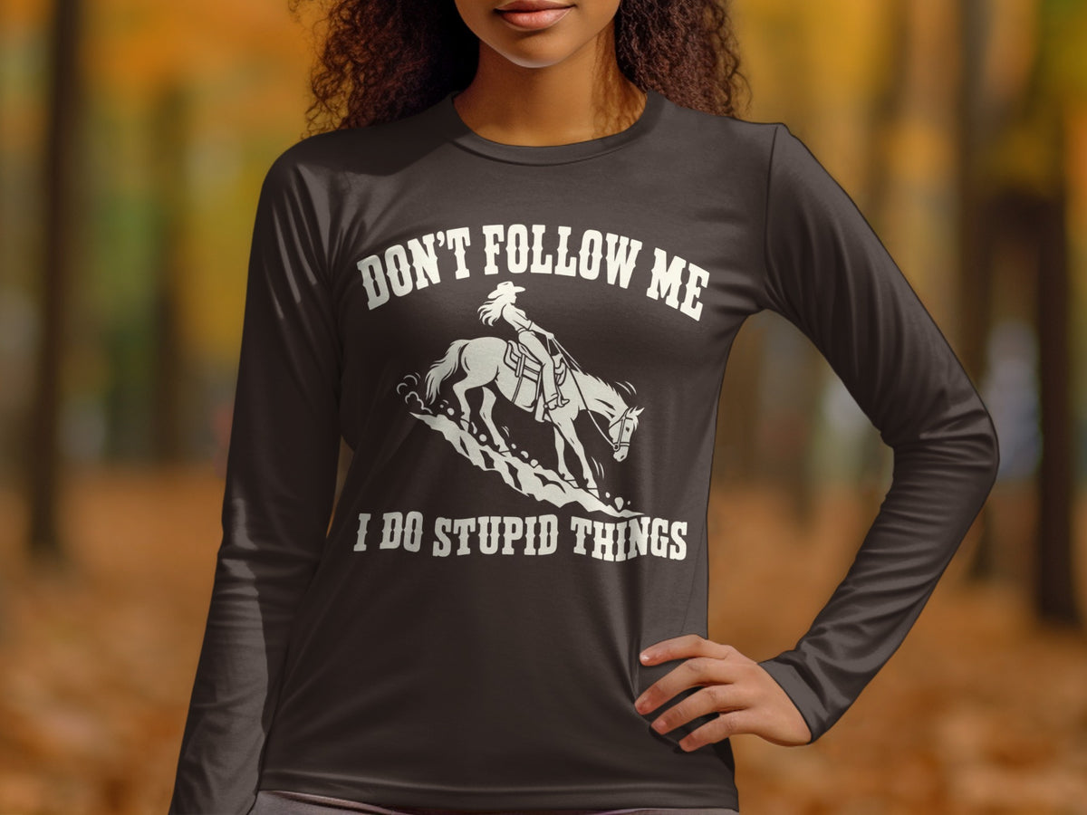 A person wearing a dark grey long-sleeve shirt with white text reading DON'T FOLLOW ME I DO STUPID THINGS and an illustration of a cowgirl riding a horse down a slope.