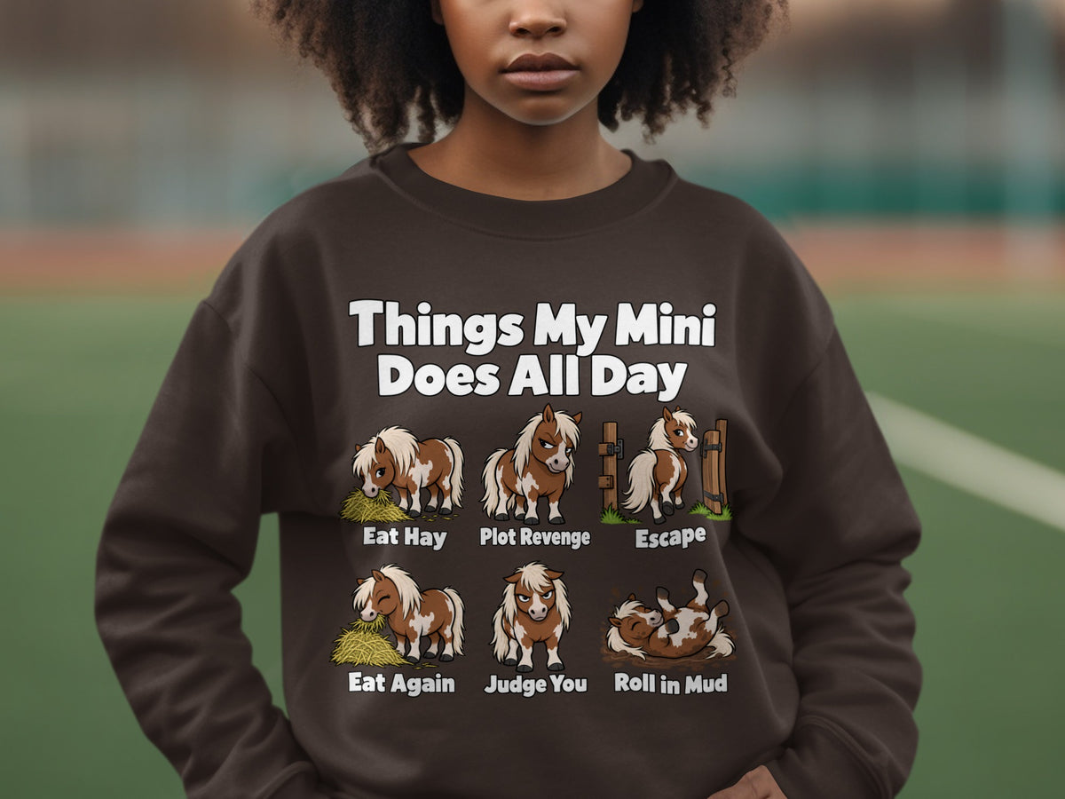 A person wearing a dark brown crewneck sweatshirt featuring illustrations and text about miniature horse activities.