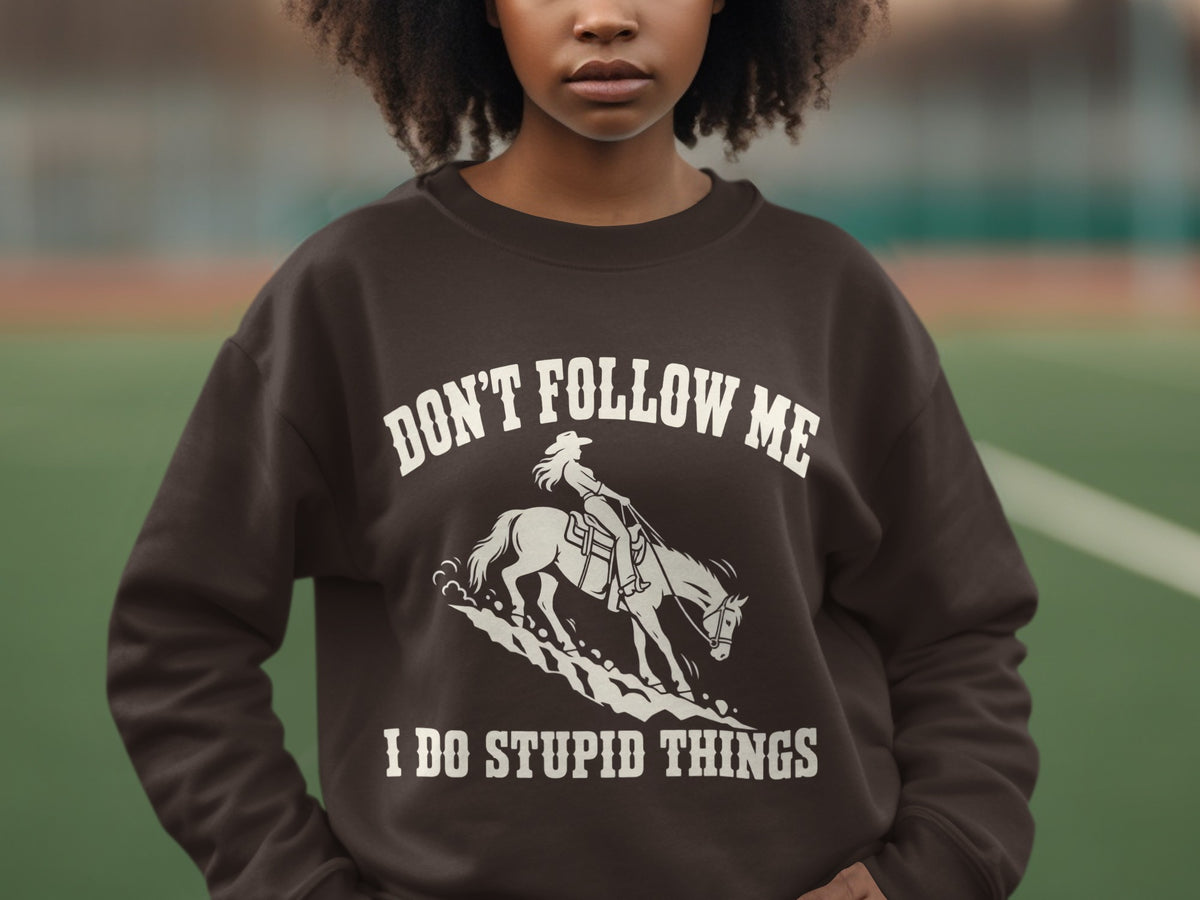 A person wearing a brown crewneck sweatshirt featuring a graphic of a cowgirl riding a horse and the text DON'T FOLLOW ME I DO STUPID THINGS.