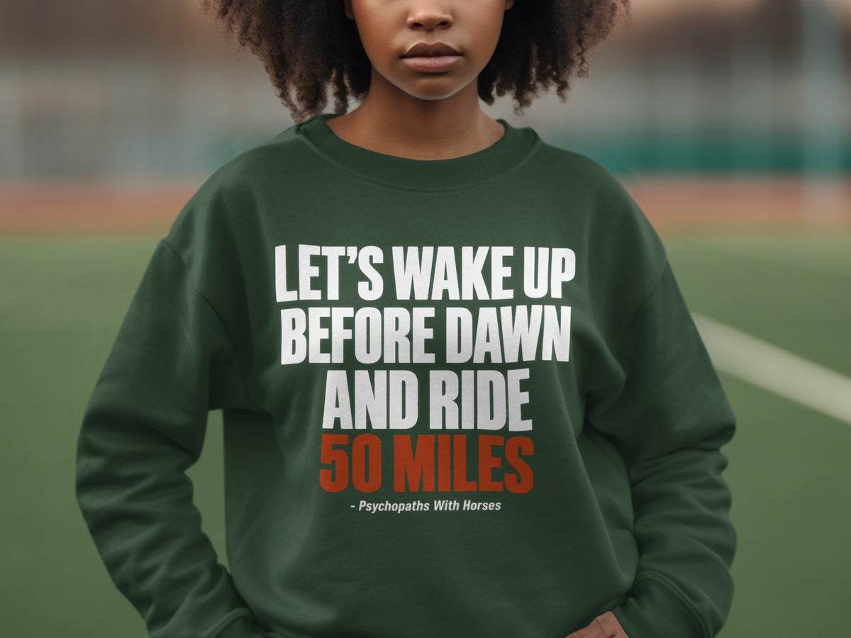 A person wearing a dark green sweatshirt printed with the text "LET'S WAKE UP BEFORE DAWN AND RIDE 50 MILES" in white and orange.